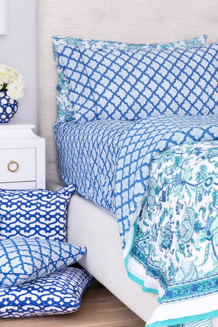 Close-up of a bed with blue and white patterned pillows and matching block-printed cotton sheets from the Jemina Sheet Set by Roller Rabbit, featuring an intricate geometric design; the headboard has a subtle white floral pattern.