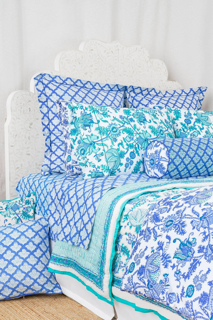 Close-up of a bed with blue and white patterned pillows and matching block-printed cotton sheets from the Jemina Sheet Set by Roller Rabbit, featuring an intricate geometric design; the headboard has a subtle white floral pattern.