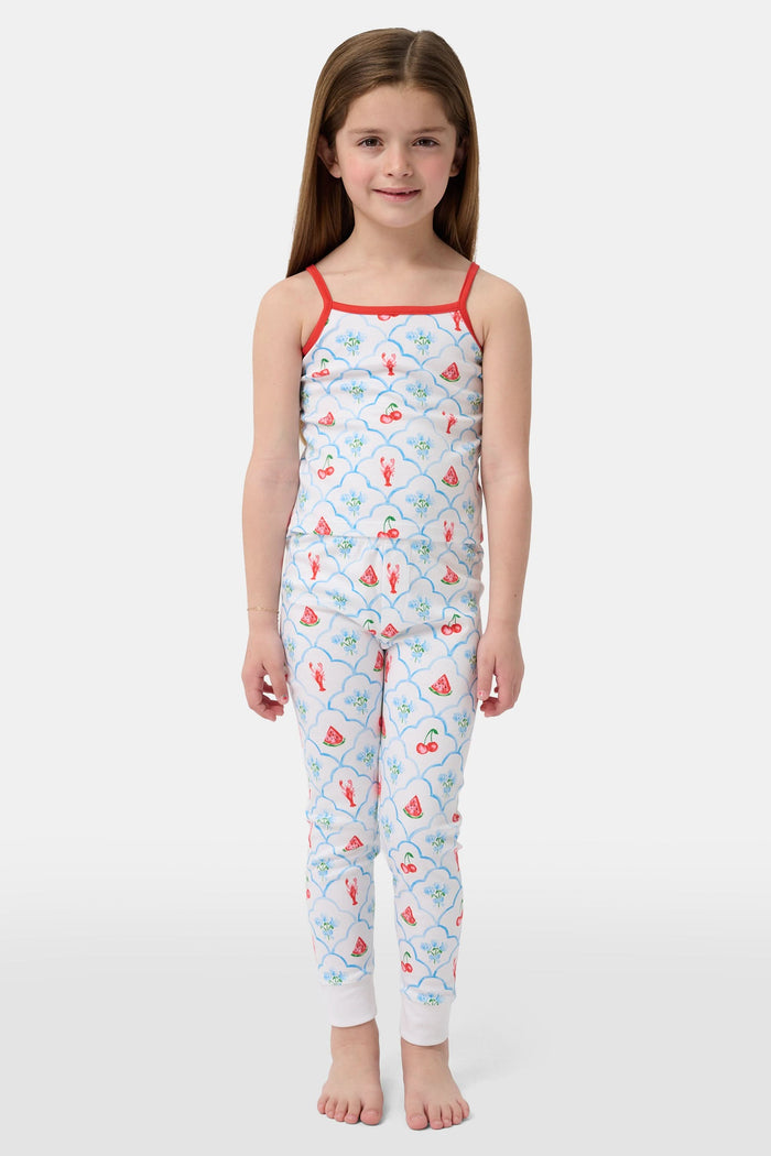 A young girl stands barefoot, smiling against a light background. She wears the Roller Rabbit Kids 4th of Jemina Noa Set—matching white Pima Cotton pajamas with red trim and a colorful flowers, butterflies, and birds print.