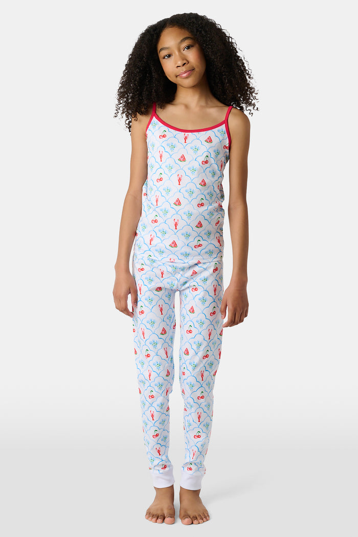 A young girl stands barefoot, smiling against a light background. She wears the Roller Rabbit Kids 4th of Jemina Noa Set—matching white Pima Cotton pajamas with red trim and a colorful flowers, butterflies, and birds print.