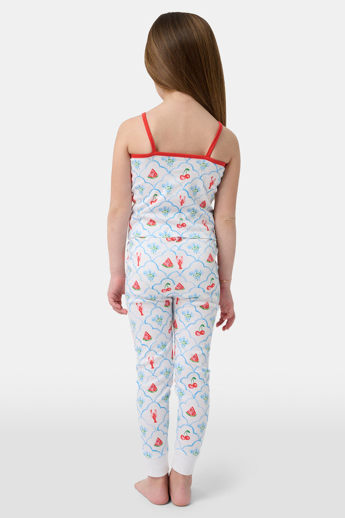 A young girl stands barefoot, smiling against a light background. She wears the Roller Rabbit Kids 4th of Jemina Noa Set—matching white Pima Cotton pajamas with red trim and a colorful flowers, butterflies, and birds print.