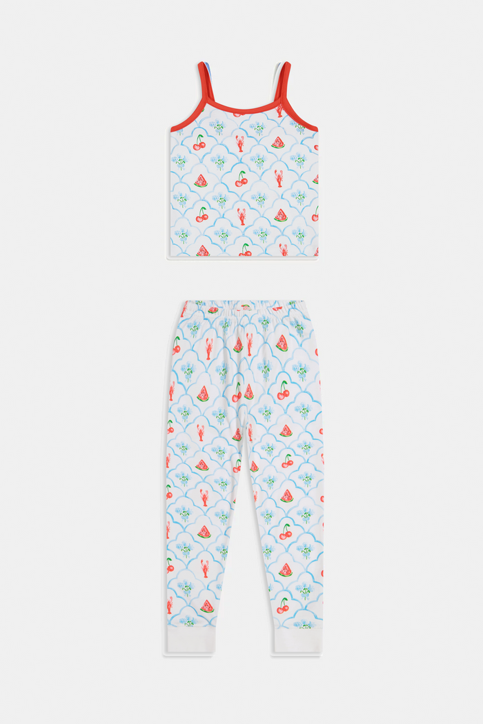 A young girl stands barefoot, smiling against a light background. She wears the Roller Rabbit Kids 4th of Jemina Noa Set—matching white Pima Cotton pajamas with red trim and a colorful flowers, butterflies, and birds print.