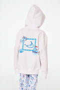 A child is wearing the Roller Rabbit Kids ACK Zip Up hoodie in white, crafted from lightweight French terry and showcasing blue and red nautical designs—including an anchor, shell, and sailboat—on the back. The child is paired with patterned white pants and is turned away to highlight the hoodie’s island-inspired details.