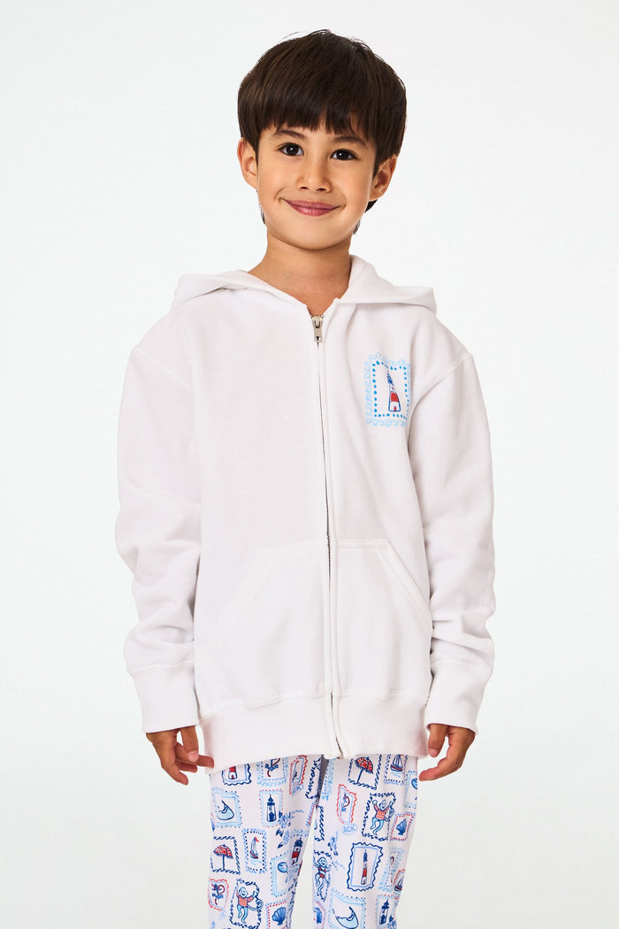 A child is wearing the Roller Rabbit Kids ACK Zip Up hoodie in white, crafted from lightweight French terry and showcasing blue and red nautical designs—including an anchor, shell, and sailboat—on the back. The child is paired with patterned white pants and is turned away to highlight the hoodie’s island-inspired details.