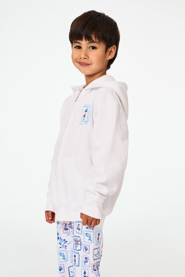A child is wearing the Roller Rabbit Kids ACK Zip Up hoodie in white, crafted from lightweight French terry and showcasing blue and red nautical designs—including an anchor, shell, and sailboat—on the back. The child is paired with patterned white pants and is turned away to highlight the hoodie’s island-inspired details.