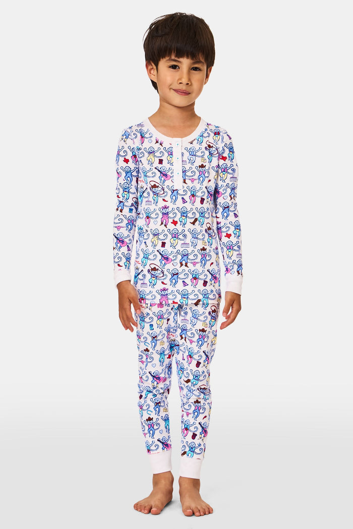 A young child stands smiling, wearing the Roller Rabbit Kids Austin Monkey Pajamas—a super soft pajama set with colorful, long sleeves and a playful cartoon monkey pattern on a white background.