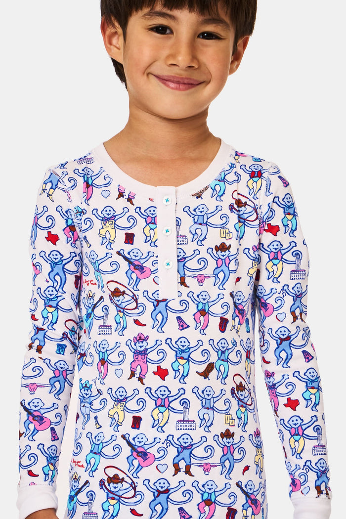 A young child stands smiling, wearing the Roller Rabbit Kids Austin Monkey Pajamas—a super soft pajama set with colorful, long sleeves and a playful cartoon monkey pattern on a white background.