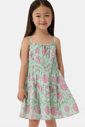 A young girl with long brown hair smiles, wearing the Roller Rabbit Kids Camellia Block Karla Dress, a sleeveless mint green cotton dress with pink floral patterns. She stands with one hand by her side and the other holding her dress.