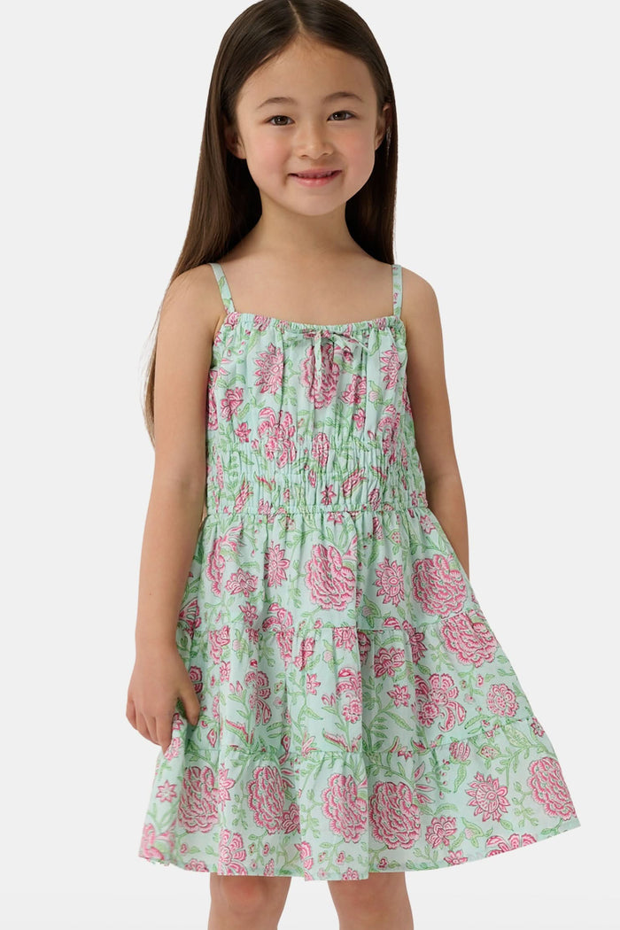 A young girl with long brown hair smiles, wearing the Roller Rabbit Kids Camellia Block Karla Dress, a sleeveless mint green cotton dress with pink floral patterns. She stands with one hand by her side and the other holding her dress.