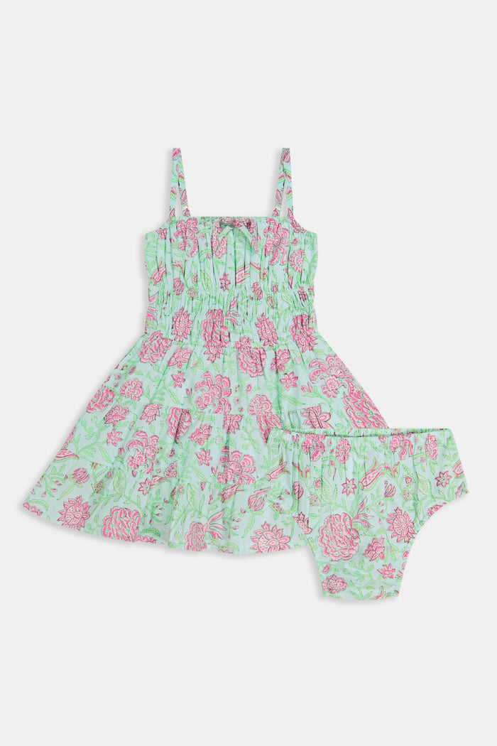 A young girl with long brown hair smiles, wearing the Roller Rabbit Kids Camellia Block Karla Dress, a sleeveless mint green cotton dress with pink floral patterns. She stands with one hand by her side and the other holding her dress.