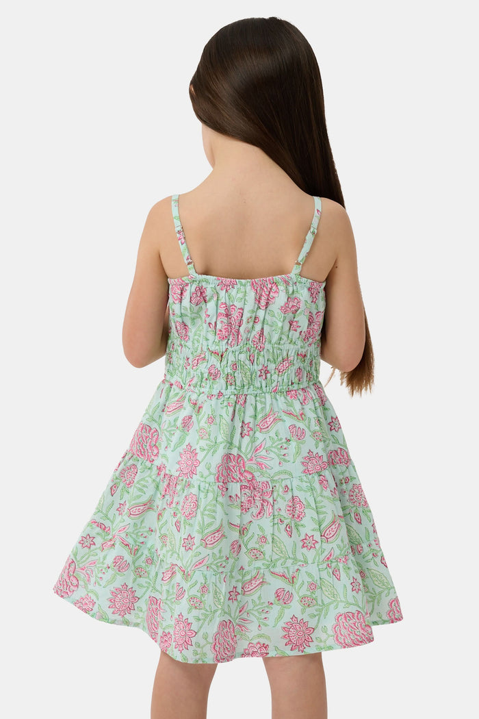 A young girl with long brown hair smiles, wearing the Roller Rabbit Kids Camellia Block Karla Dress, a sleeveless mint green cotton dress with pink floral patterns. She stands with one hand by her side and the other holding her dress.