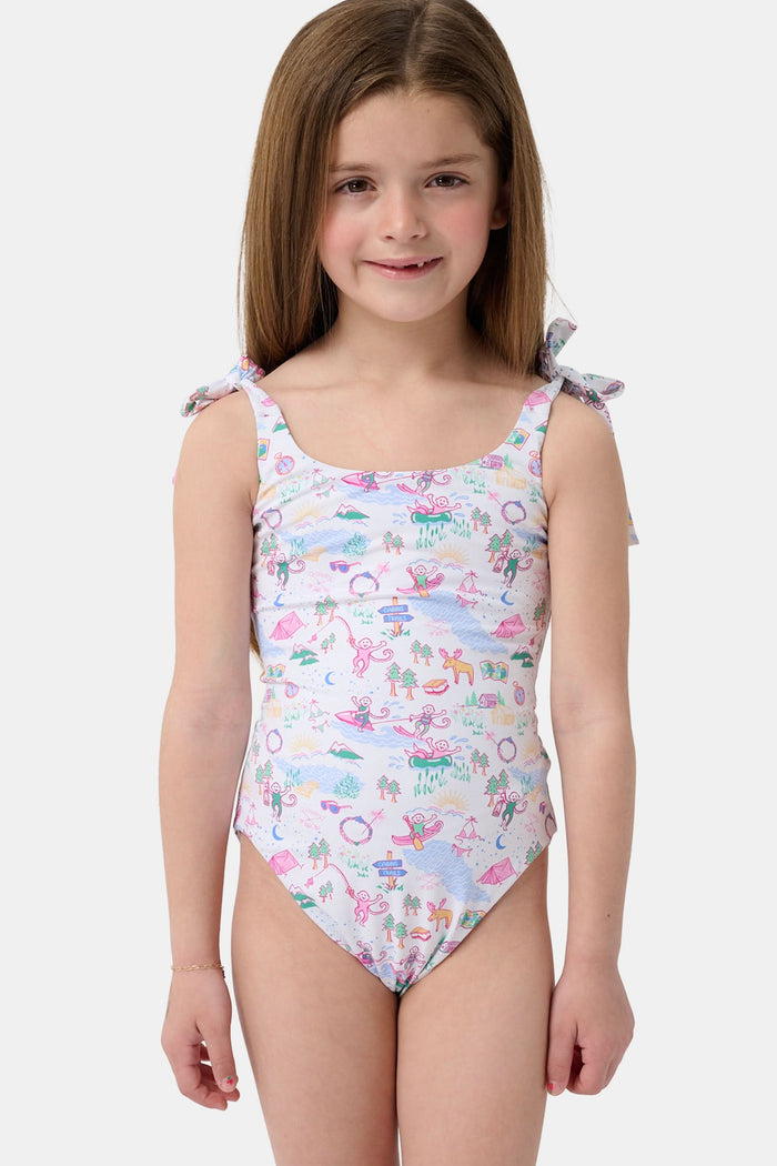 A young girl with straight brown hair smiles in the Roller Rabbit Kids Camp RR Cora One Piece, a swimsuit with colorful patterns and adjustable shoulder ties. She stands against a plain, light background.