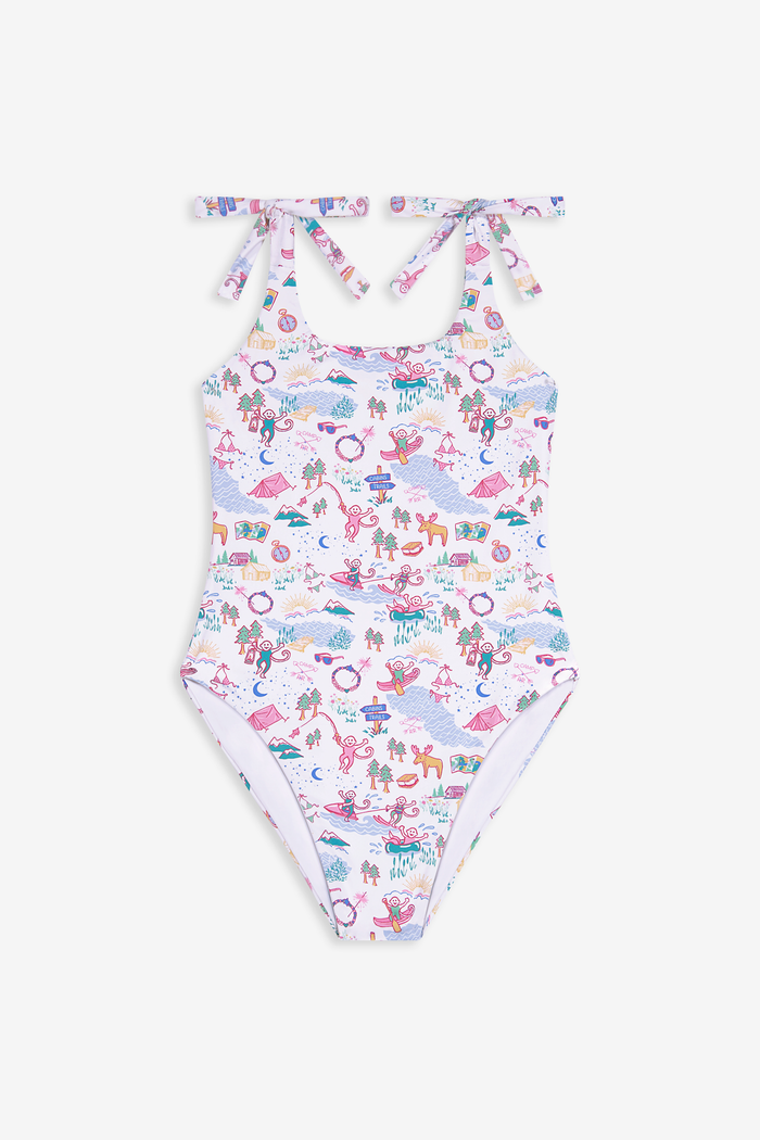 A young girl with straight brown hair smiles in the Roller Rabbit Kids Camp RR Cora One Piece, a swimsuit with colorful patterns and adjustable shoulder ties. She stands against a plain, light background.