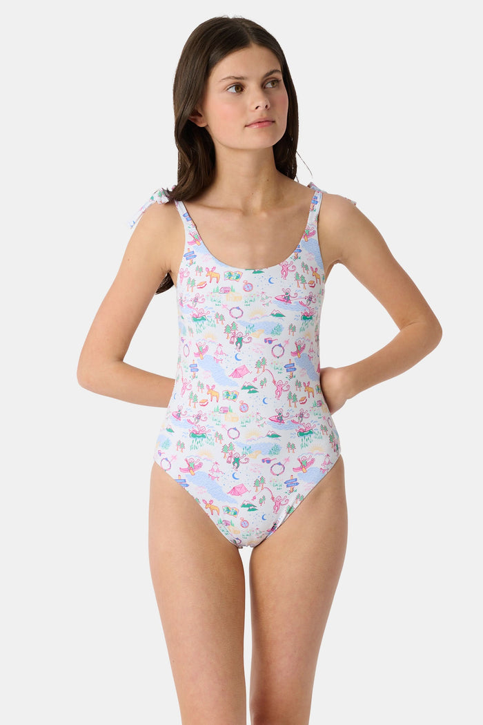 A young girl with straight brown hair smiles in the Roller Rabbit Kids Camp RR Cora One Piece, a swimsuit with colorful patterns and adjustable shoulder ties. She stands against a plain, light background.