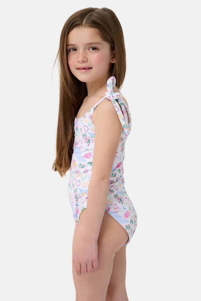 A young girl with straight brown hair smiles in the Roller Rabbit Kids Camp RR Cora One Piece, a swimsuit with colorful patterns and adjustable shoulder ties. She stands against a plain, light background.