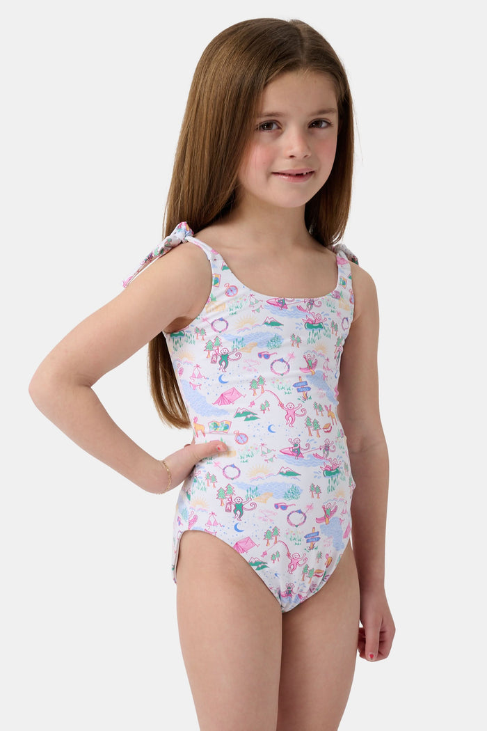 A young girl with straight brown hair smiles in the Roller Rabbit Kids Camp RR Cora One Piece, a swimsuit with colorful patterns and adjustable shoulder ties. She stands against a plain, light background.