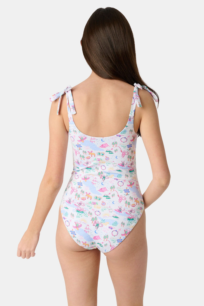 A young girl with straight brown hair smiles in the Roller Rabbit Kids Camp RR Cora One Piece, a swimsuit with colorful patterns and adjustable shoulder ties. She stands against a plain, light background.