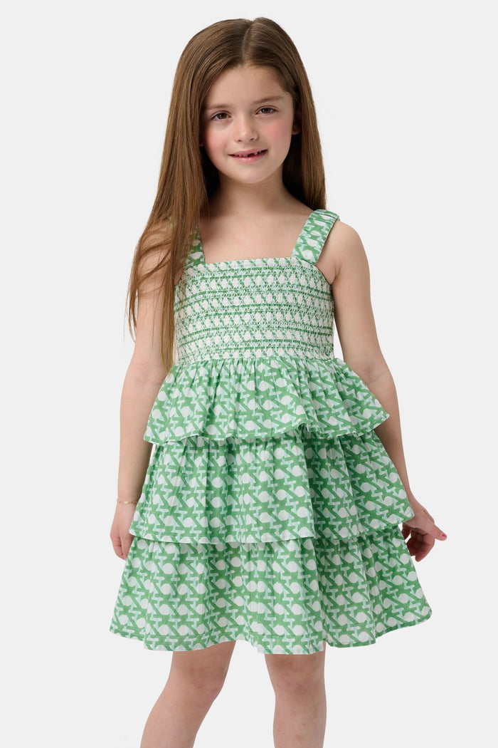 A young girl with curly hair wears the Roller Rabbit Kids Cane Kenia Dress, a green and white patterned cotton tiered dress with wide straps, standing against a plain light background.