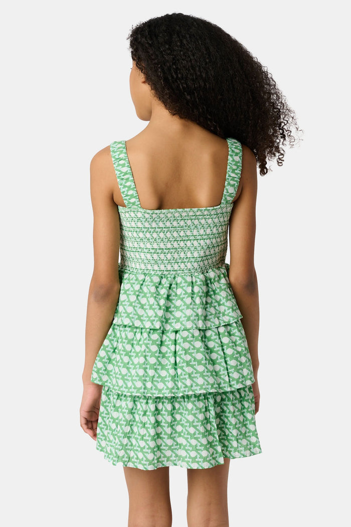 A young girl with curly hair wears the Roller Rabbit Kids Cane Kenia Dress, a green and white patterned cotton tiered dress with wide straps, standing against a plain light background.