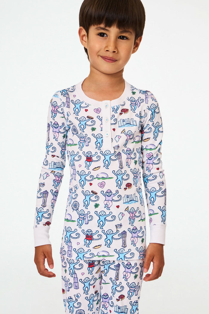 A young child with short dark hair smiles while wearing Roller Rabbit Kids Chicago Monkey Pajamas, featuring colorful cartoon monkeys, hearts, and books on a white background.