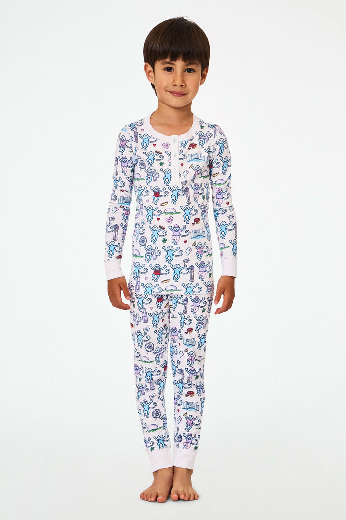 A young child with short dark hair smiles while wearing Roller Rabbit Kids Chicago Monkey Pajamas, featuring colorful cartoon monkeys, hearts, and books on a white background.