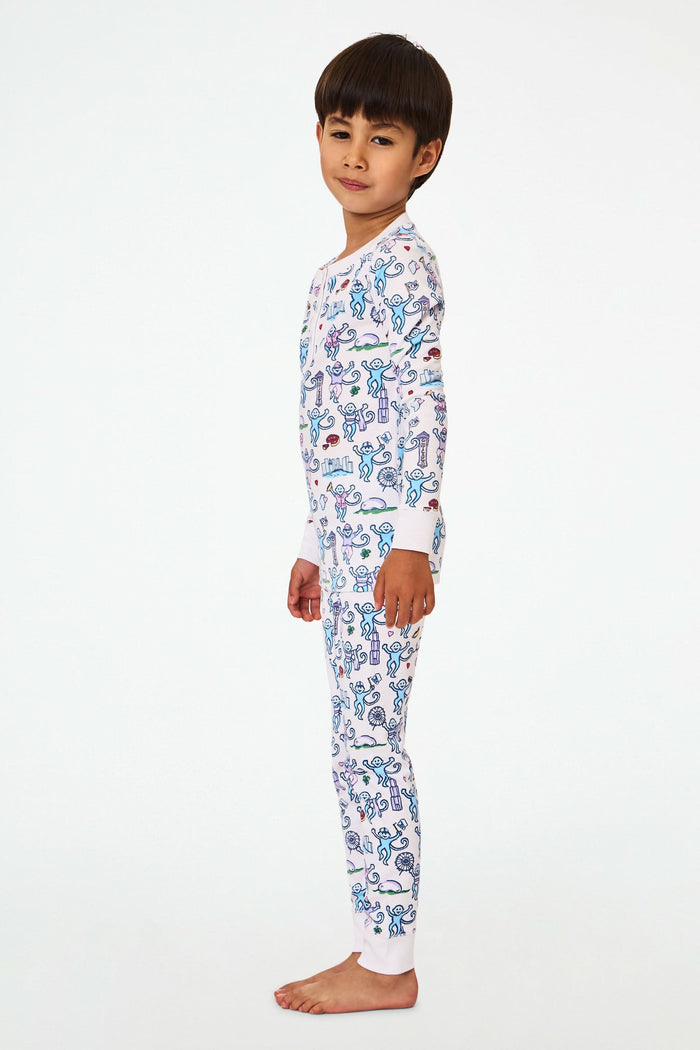 A young child with short dark hair smiles while wearing Roller Rabbit Kids Chicago Monkey Pajamas, featuring colorful cartoon monkeys, hearts, and books on a white background.