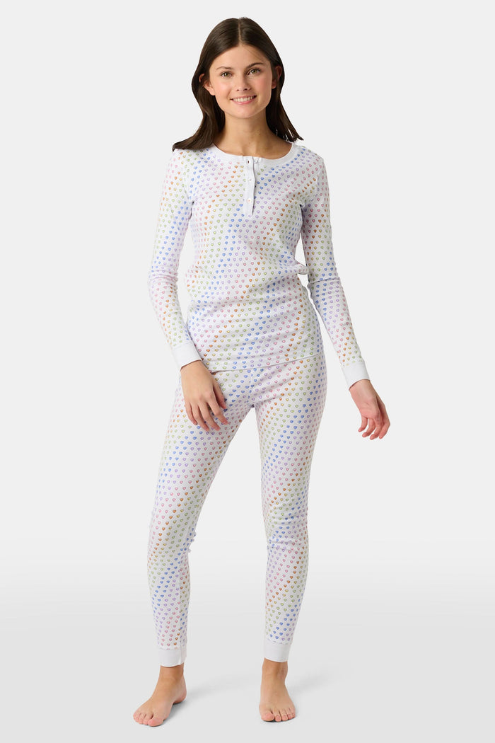 A girl wears Roller Rabbit's Kids Disco Hearts Pajamas, featuring a long-sleeve, buttoned top and matching pants with a colorful rainbow heart print on a light background.