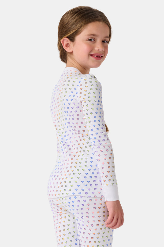 A girl wears Roller Rabbit's Kids Disco Hearts Pajamas, featuring a long-sleeve, buttoned top and matching pants with a colorful rainbow heart print on a light background.