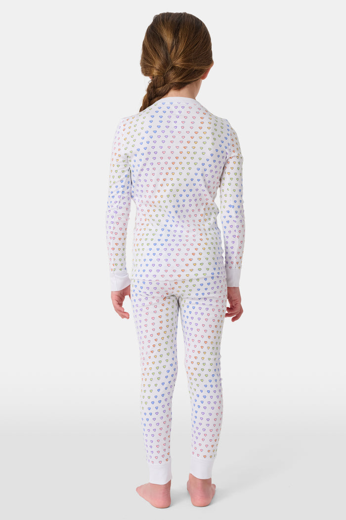 A girl wears Roller Rabbit's Kids Disco Hearts Pajamas, featuring a long-sleeve, buttoned top and matching pants with a colorful rainbow heart print on a light background.