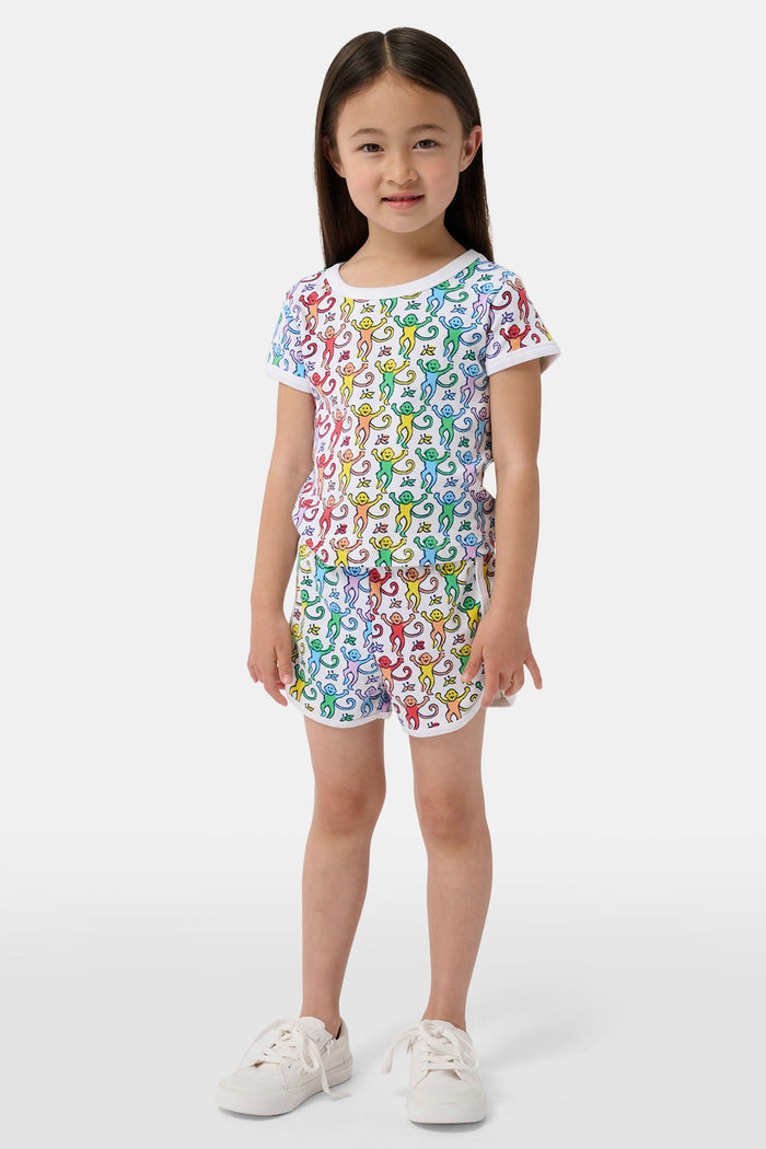 A young child smiles in the Roller Rabbit Kids Disco Monkey Playa Play Set, featuring a playful monkey print cotton shorts outfit, paired with white sneakers. The simple light background highlights this cheerful look.