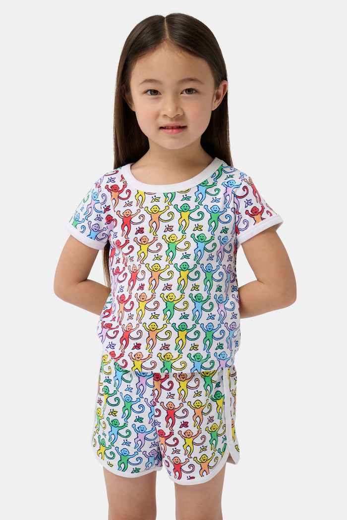 A young child smiles in the Roller Rabbit Kids Disco Monkey Playa Play Set, featuring a playful monkey print cotton shorts outfit, paired with white sneakers. The simple light background highlights this cheerful look.