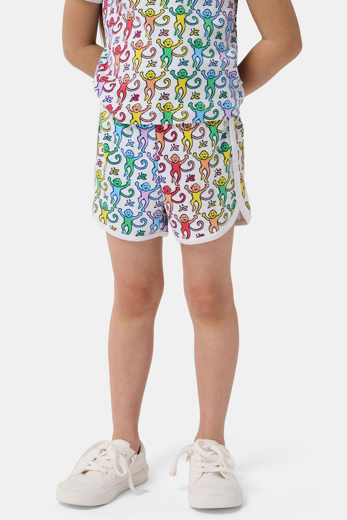 A young child smiles in the Roller Rabbit Kids Disco Monkey Playa Play Set, featuring a playful monkey print cotton shorts outfit, paired with white sneakers. The simple light background highlights this cheerful look.