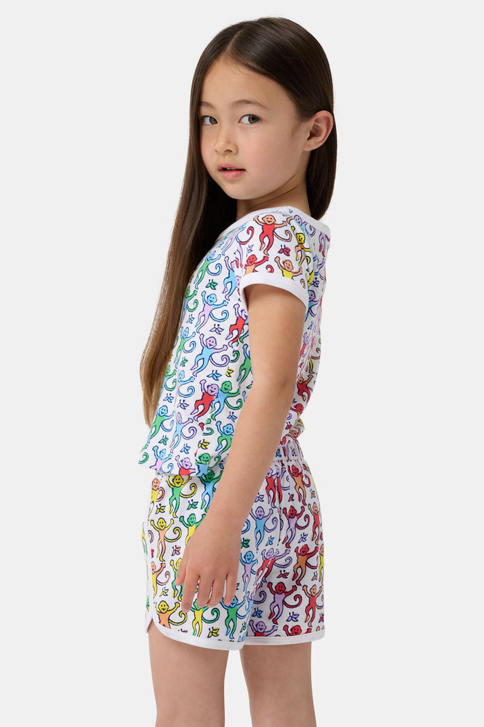 A young child smiles in the Roller Rabbit Kids Disco Monkey Playa Play Set, featuring a playful monkey print cotton shorts outfit, paired with white sneakers. The simple light background highlights this cheerful look.