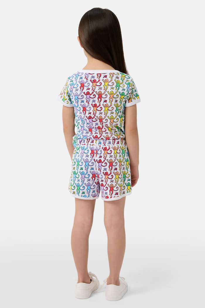 A young child smiles in the Roller Rabbit Kids Disco Monkey Playa Play Set, featuring a playful monkey print cotton shorts outfit, paired with white sneakers. The simple light background highlights this cheerful look.