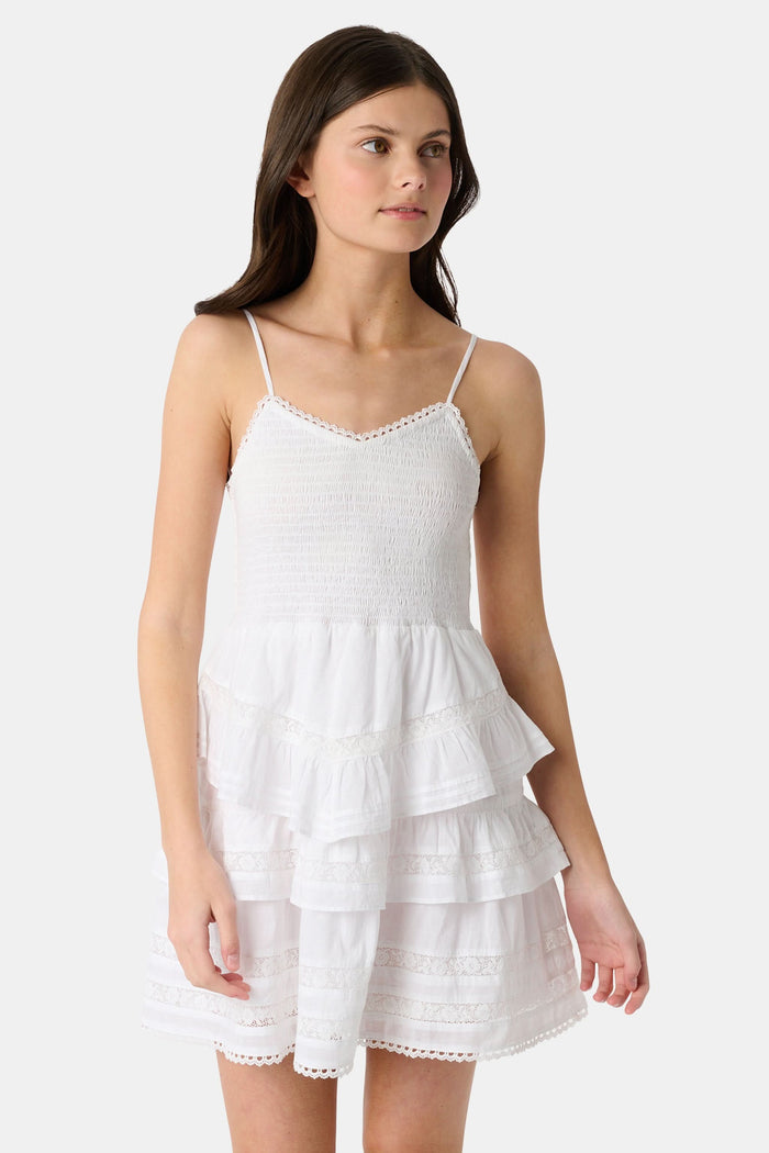 A girl with long brown hair wears the Roller Rabbit Kids Florence Lace Lennox Dress, a white sleeveless tiered cotton voile dress with thin straps and lace details, standing against a plain light grey background.