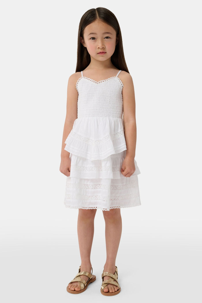 A girl with long brown hair wears the Roller Rabbit Kids Florence Lace Lennox Dress, a white sleeveless tiered cotton voile dress with thin straps and lace details, standing against a plain light grey background.