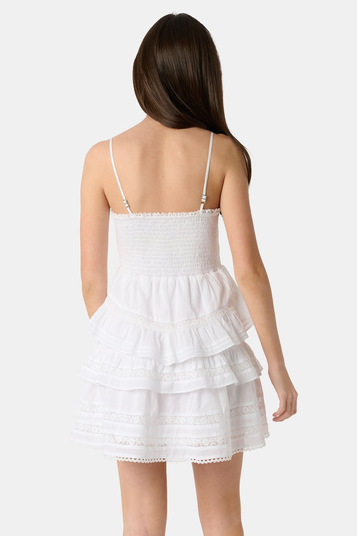 A girl with long brown hair wears the Roller Rabbit Kids Florence Lace Lennox Dress, a white sleeveless tiered cotton voile dress with thin straps and lace details, standing against a plain light grey background.