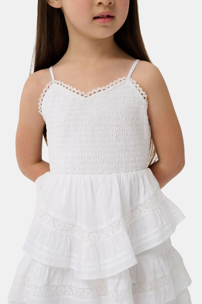 A girl with long brown hair wears the Roller Rabbit Kids Florence Lace Lennox Dress, a white sleeveless tiered cotton voile dress with thin straps and lace details, standing against a plain light grey background.