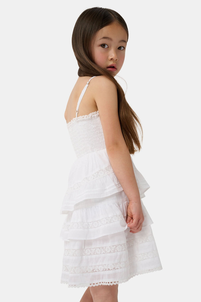 A girl with long brown hair wears the Roller Rabbit Kids Florence Lace Lennox Dress, a white sleeveless tiered cotton voile dress with thin straps and lace details, standing against a plain light grey background.