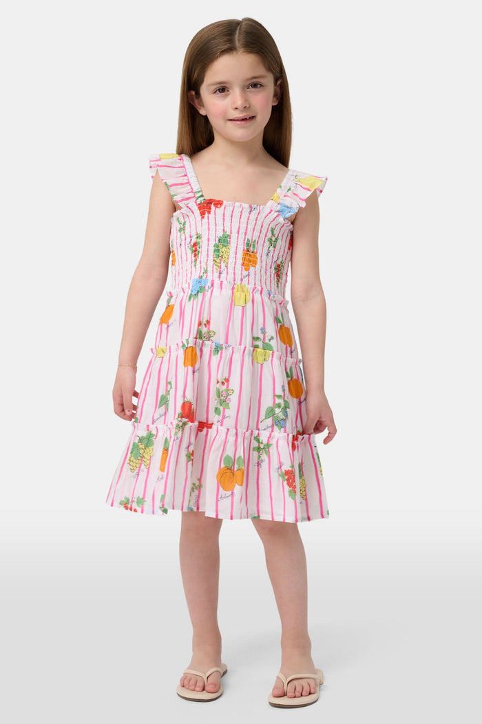 A young girl with straight brown hair wears the Roller Rabbit Kids Fruits D'Ete Ceri Dress, a pink and white smocked sundress with ruffled sleeves, and white sandals, standing and smiling against a light gray background.