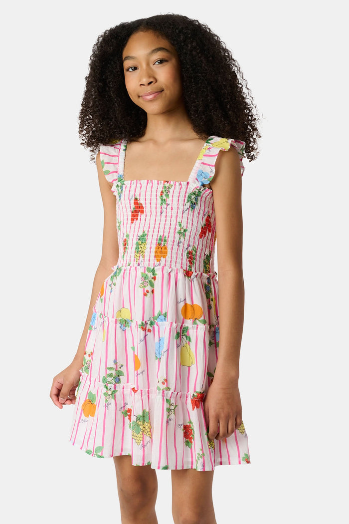 A young girl with straight brown hair wears the Roller Rabbit Kids Fruits D'Ete Ceri Dress, a pink and white smocked sundress with ruffled sleeves, and white sandals, standing and smiling against a light gray background.