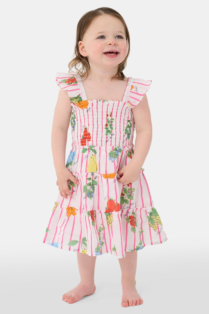 A young girl with straight brown hair wears the Roller Rabbit Kids Fruits D'Ete Ceri Dress, a pink and white smocked sundress with ruffled sleeves, and white sandals, standing and smiling against a light gray background.