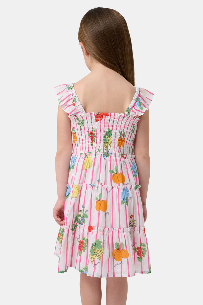 A young girl with straight brown hair wears the Roller Rabbit Kids Fruits D'Ete Ceri Dress, a pink and white smocked sundress with ruffled sleeves, and white sandals, standing and smiling against a light gray background.