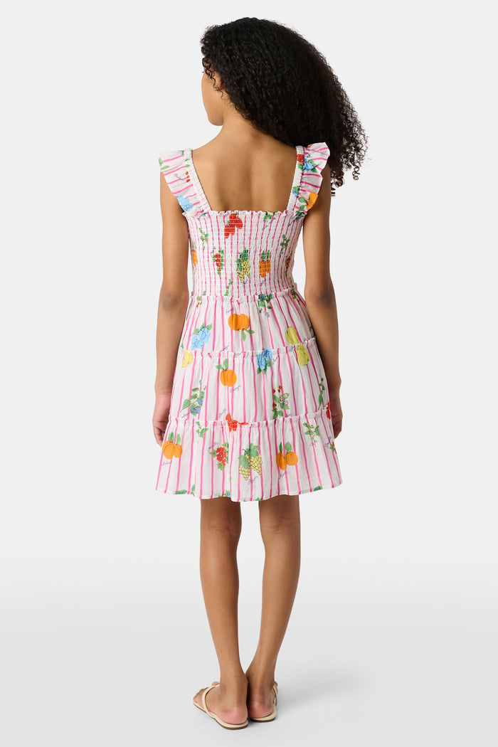 A young girl with straight brown hair wears the Roller Rabbit Kids Fruits D'Ete Ceri Dress, a pink and white smocked sundress with ruffled sleeves, and white sandals, standing and smiling against a light gray background.