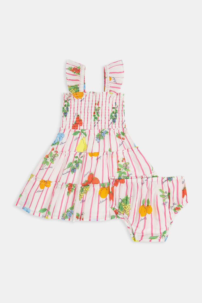 A young girl with straight brown hair wears the Roller Rabbit Kids Fruits D'Ete Ceri Dress, a pink and white smocked sundress with ruffled sleeves, and white sandals, standing and smiling against a light gray background.