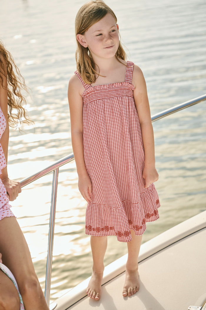 A young girl in the Roller Rabbit Kids Gingham Teagan Dress with an embroidered hem stands barefoot on a boat deck by the water, sunlight shimmering on the surface.