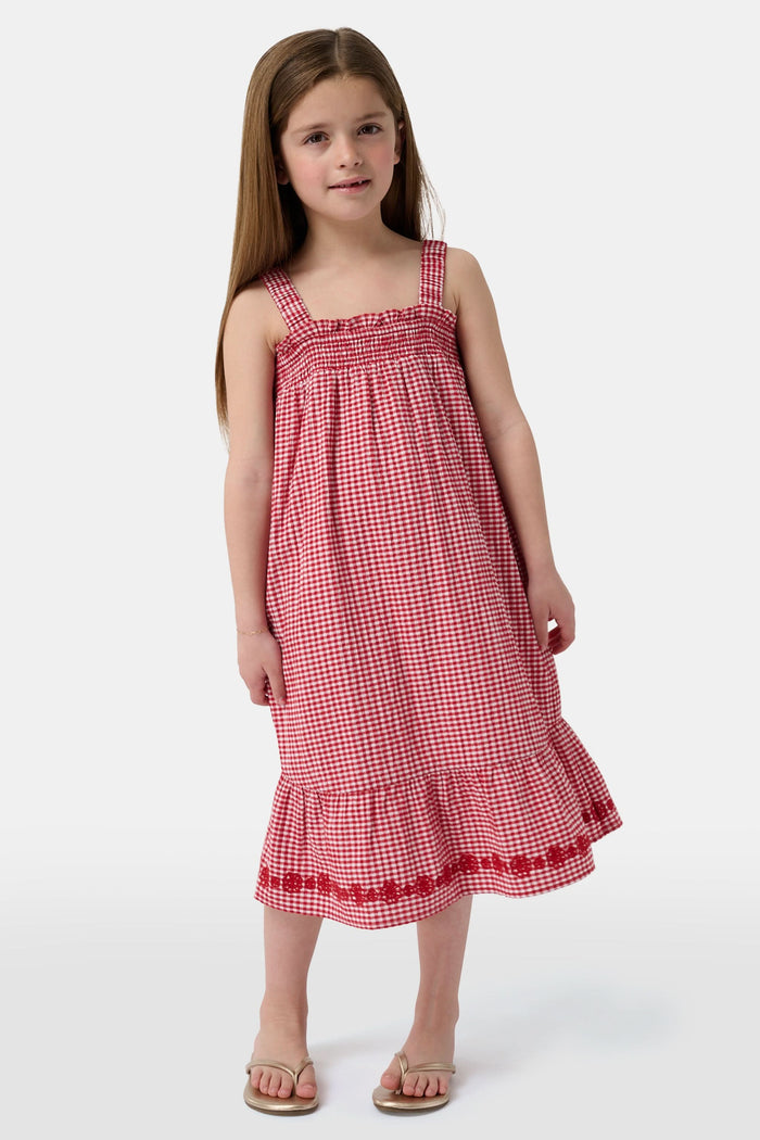 A young girl in the Roller Rabbit Kids Gingham Teagan Dress with an embroidered hem stands barefoot on a boat deck by the water, sunlight shimmering on the surface.