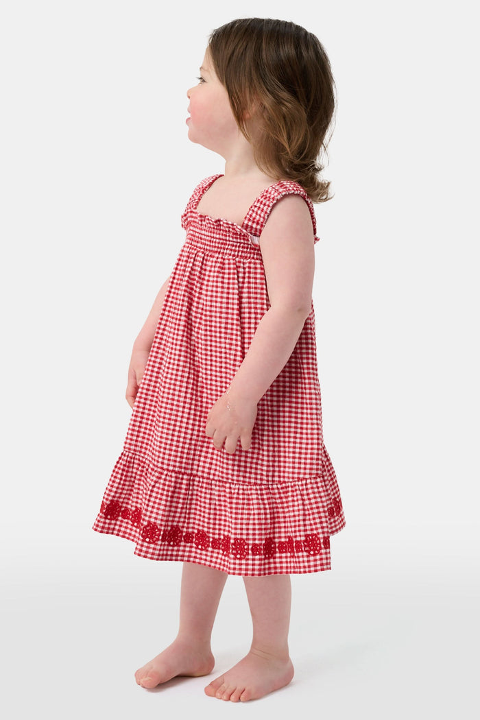A young girl in the Roller Rabbit Kids Gingham Teagan Dress with an embroidered hem stands barefoot on a boat deck by the water, sunlight shimmering on the surface.