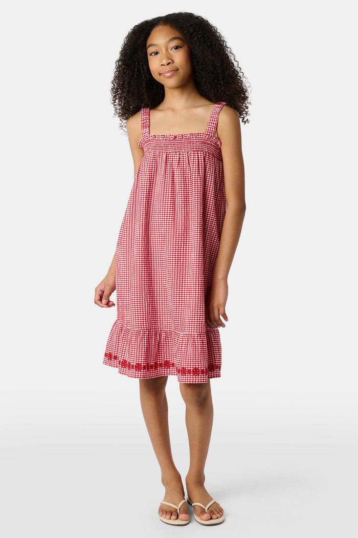 A young girl in the Roller Rabbit Kids Gingham Teagan Dress with an embroidered hem stands barefoot on a boat deck by the water, sunlight shimmering on the surface.