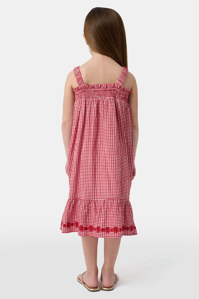 A young girl in the Roller Rabbit Kids Gingham Teagan Dress with an embroidered hem stands barefoot on a boat deck by the water, sunlight shimmering on the surface.